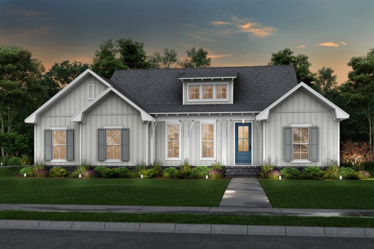 A modern single-story house with a gray exterior featuring board and batten siding. The home has a central blue front door with a small porch, flanked by two symmetrical sets of windows with decorative shutters. The roof is a dark gray shingle, and there are subtle gables on either side of the house. The front yard is neatly landscaped with a variety of shrubs and plants, and a paved pathway leads to the entrance. The setting is during twilight, creating a warm, inviting glow from the interior lights visible through the windows.