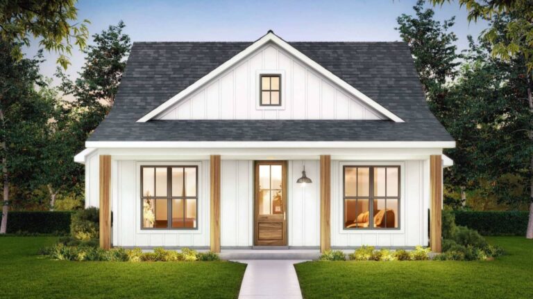 A front view of a small, modern farmhouse-style home with a cozy and charming design. The exterior features white board and batten siding, a dark shingled roof, and natural wood accents on the columns flanking the front door. The house has a covered front porch, leading to a wooden front door with glass panels. Large windows on either side of the door allow ample natural light into the home. The front yard is landscaped with a lush green lawn and small shrubs, with mature trees surrounding the home, all set against a clear evening sky.